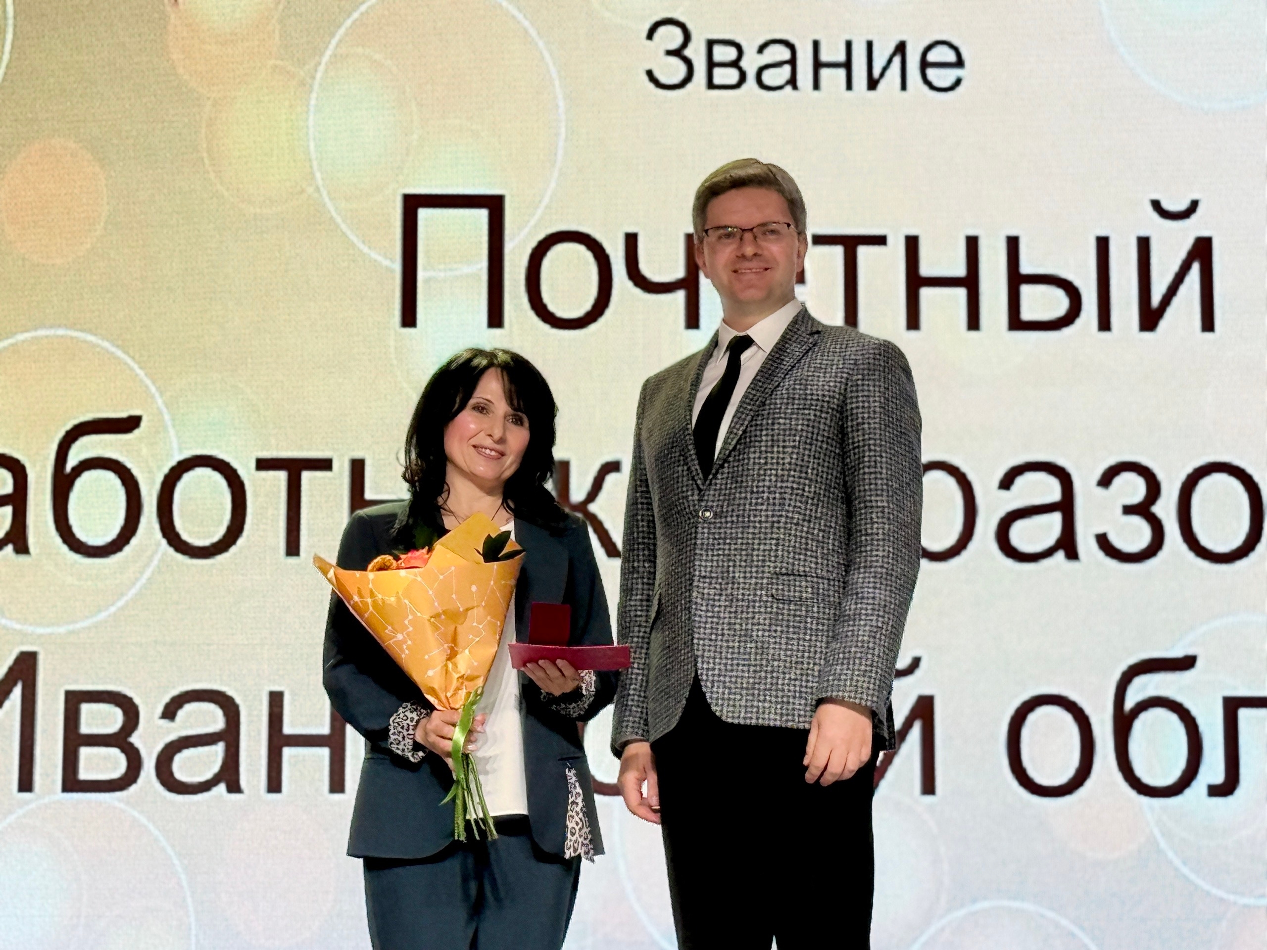 Женщина с букетом и футляром награды стоит на сцене рядом с мужчиной; на фоне проекционный экран с крупными словами «Звание», «Почетный» и частью слова «работник/Иван…»