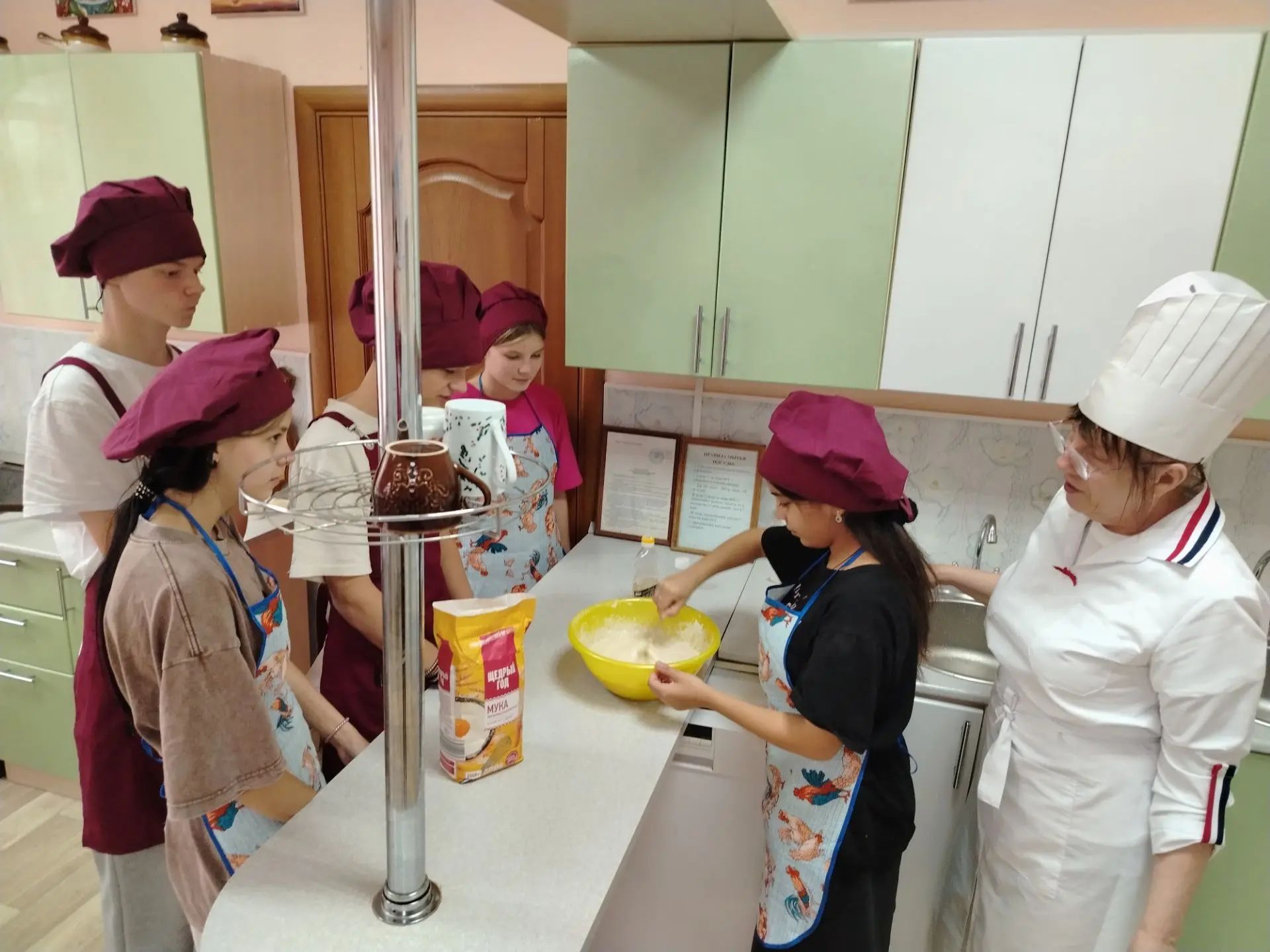 Воспитанники в поварских колпаках и фартуках замешивают тесто в кухне под руководством взрослого в поварской форме