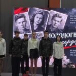 Группа школьников/участников на сцене перед большим экраном с изображениями молодогвардейцев и надписями о «Молодой гвардии».