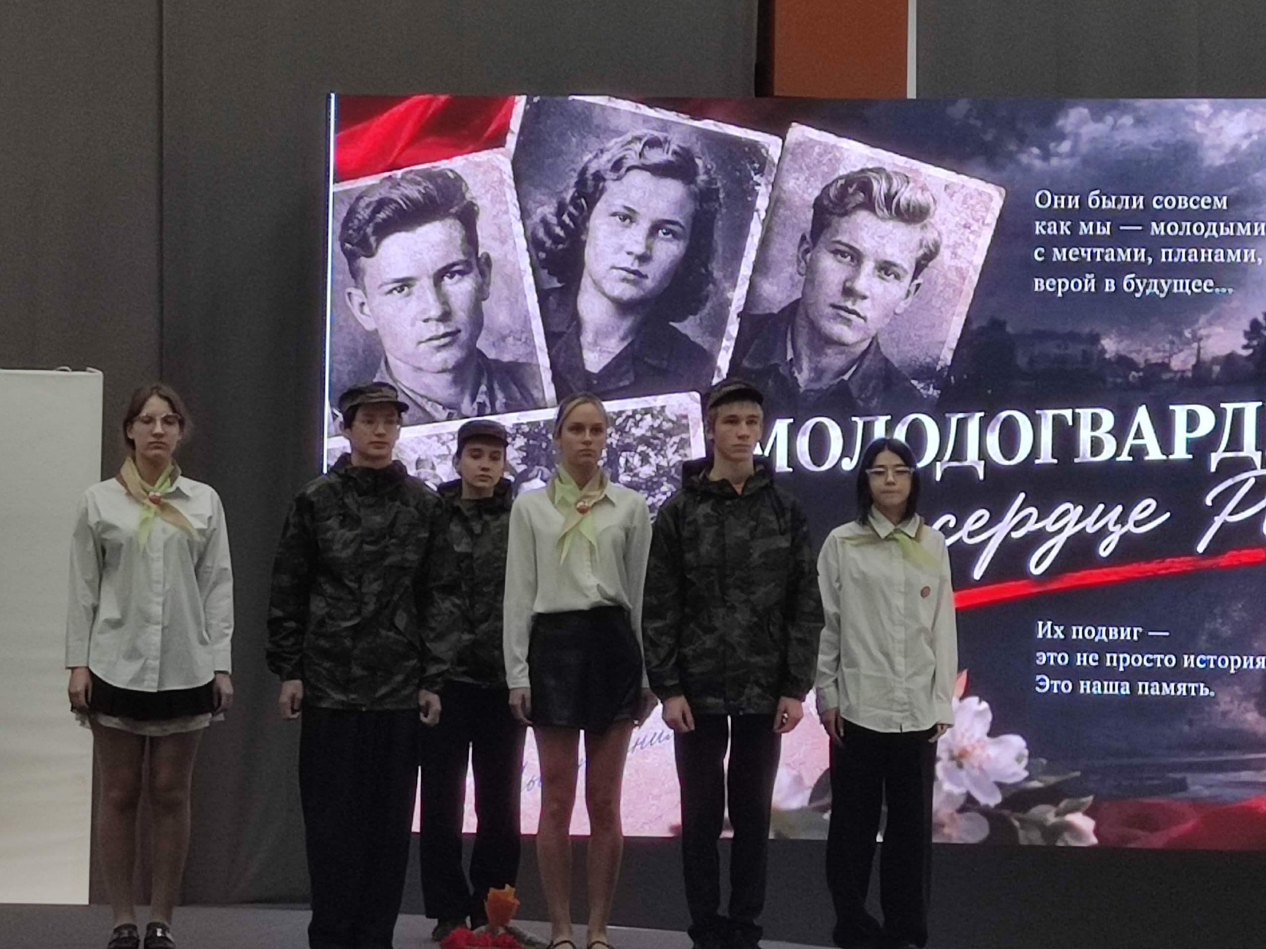 Группа школьников/участников на сцене перед большим экраном с изображениями молодогвардейцев и надписями о «Молодой гвардии».