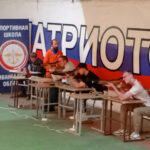 Несколько стрелков в спортивном тире сидят за столами и стреляют из пневматических винтовок на фоне большого баннера с триколором и надписью «ПАТРИОТ» и стенда «СПОРТИВНАЯ ШКОЛА Ивановской обл.»