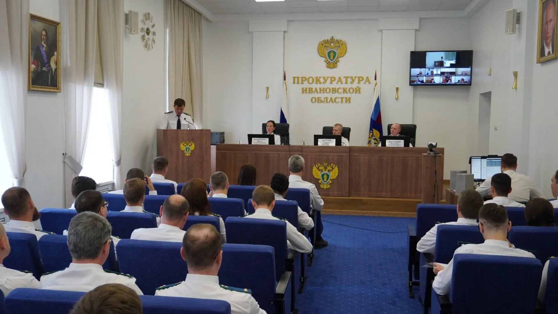 Зал заседаний прокуратуры с трибуной, гербом и надписью «Прокуратура Ивановской области», люди в светлой форме в аудитории.