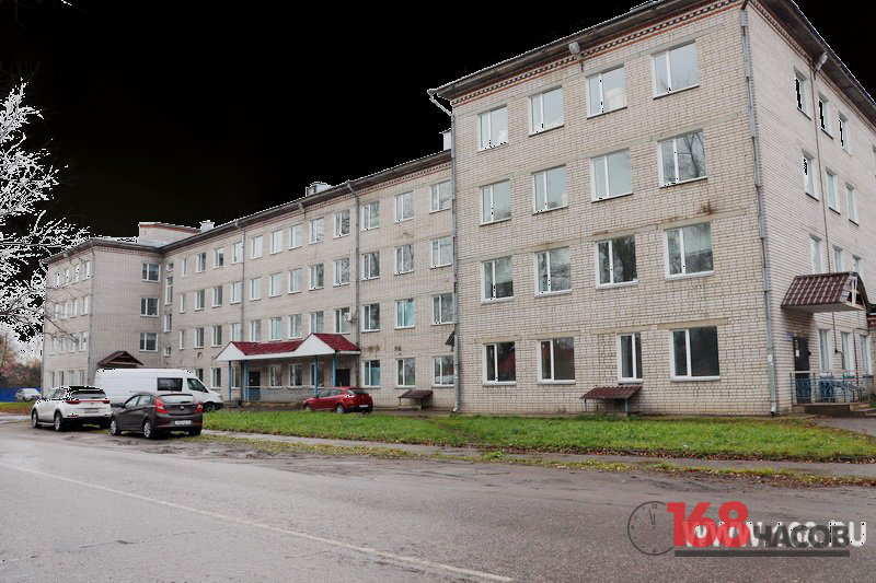 Многоэтажное кирпичное здание медицинского учреждения с припаркованными автомобилями и логотипом издания в углу