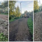 Нерегулируемая грунтовая/гравийная дорога в зелёной сельской застройке: дорожная техника (экскаватор-погрузчик), заборы и опоры линий электропередачи.