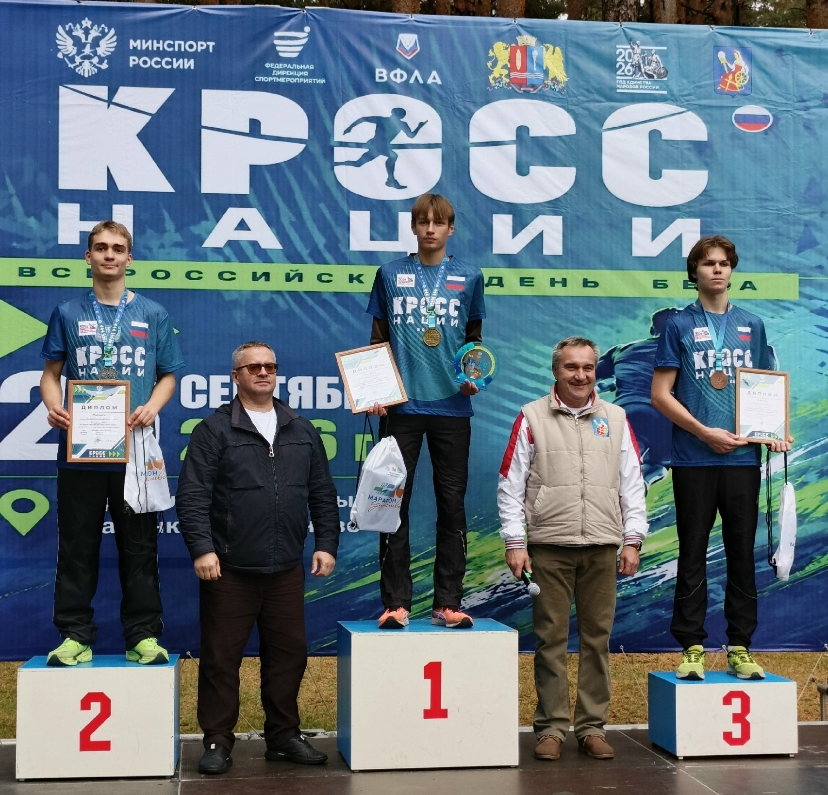 Пьедестал почёта с тремя призёрами на фоне большого баннера «Кросс нации»: медали, дипломы и номера подиумов 1–2–3.