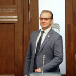 Мужчина в деловом костюме и очках стоит в кабинете у деревянной двери с табличкой «ЗАМЕСТИТЕЛЬ ПРЕДСЕДАТЕЛЯ ГОСУДАРСТВЕННОЙ ДУМЫ»; на стекле виден логотип «ЕДИНАЯ РОССИЯ».