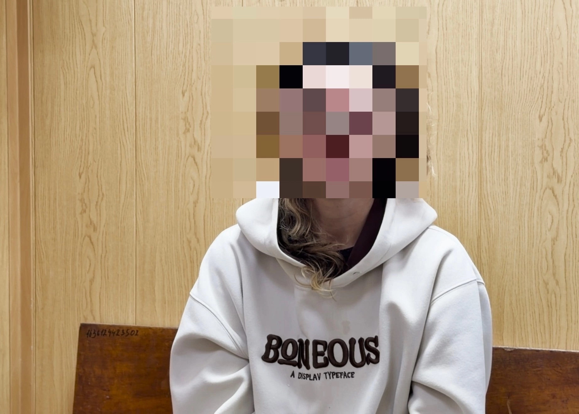 сидящая женщина в светлом худи на фоне деревянной панели; лицо замазано/пикселизировано
