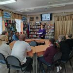 Группа людей в читальном зале библиотеки слушает женщину-ведущую на фоне книжных полок и экрана с биографическими слайдами.