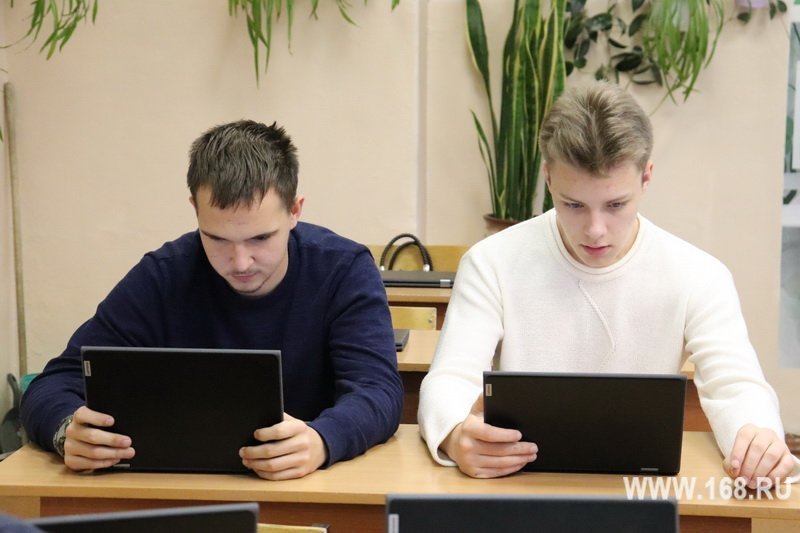 Двое молодых людей за партами работают на планшетах/ноутбуках в учебном классе/кабинете