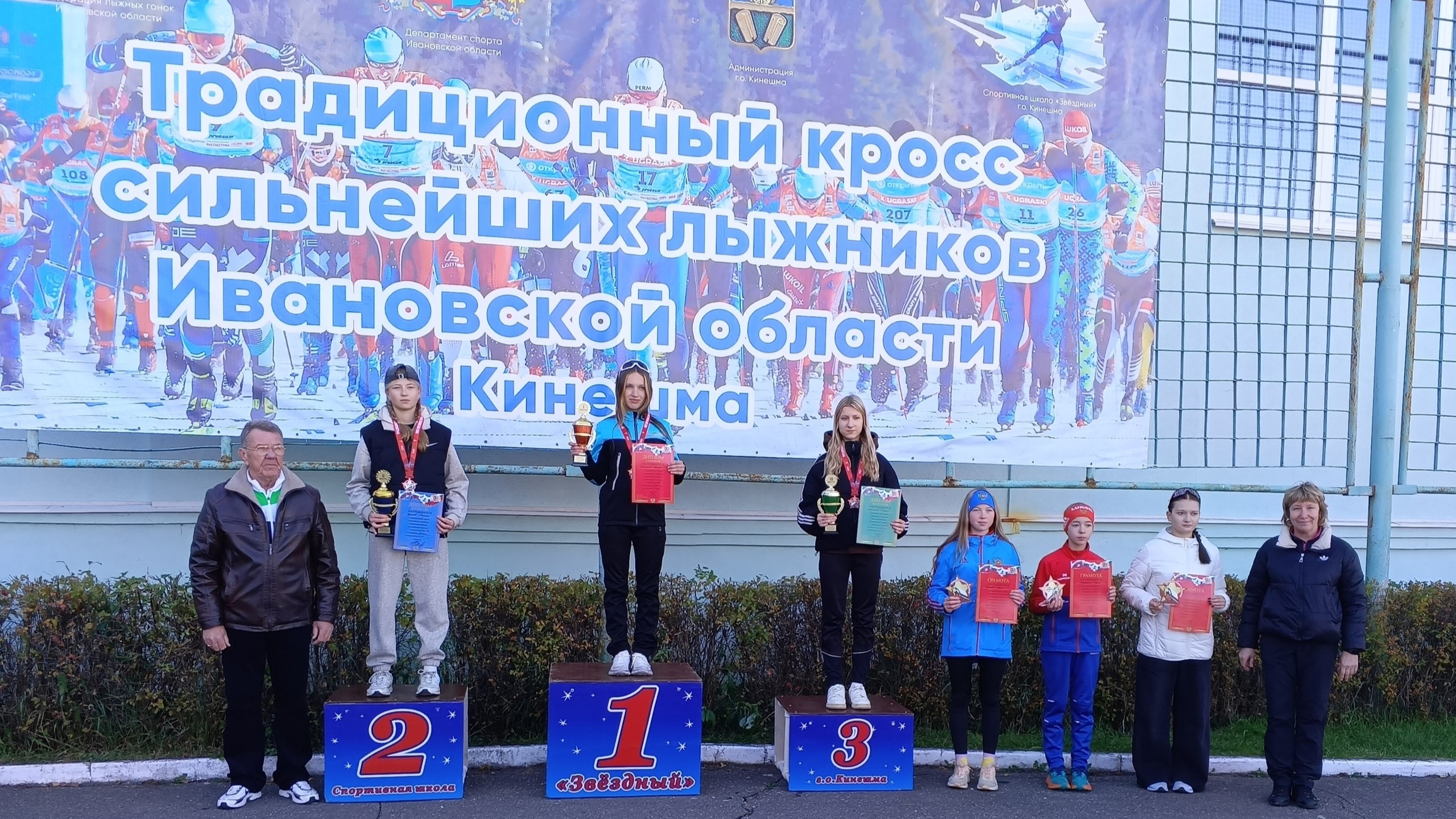 Церемония награждения юных лыжников на пьедестале с кубками и дипломами на фоне большого баннера «Традиционный кросс сильнейших лыжников Ивановской области — Кинешма»