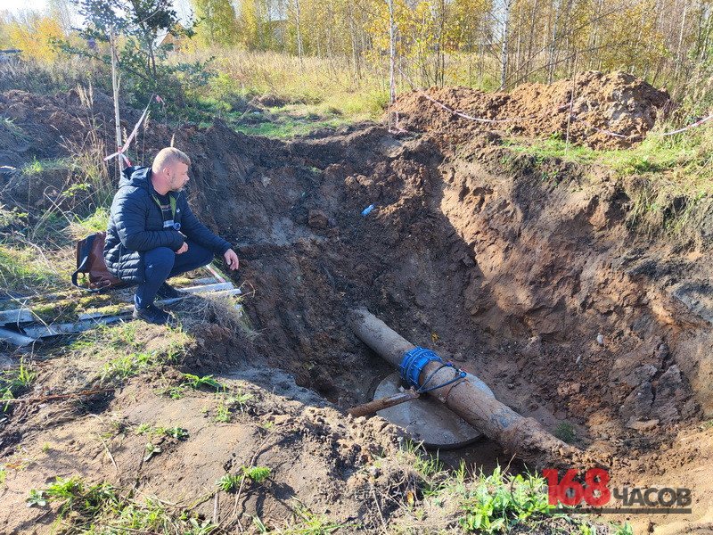 траншея с проложенной трубой и муфтой для канализации; рабочий осматривает раскоп; ленты ограждения и растительность на заднем плане