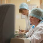 Две медицинские работницы в белых халатах и одноразовых шапочках в амбулаторном кабинете у компьютера; на заднем плане полки с папками медкарт.