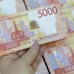 Рука держит купюру 5000 рублей на фоне нескольких таких купюр