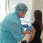 медицинская работница в голубой форме и шапочке делает укол (прививку) в плечо молодой женщине в процедурном кабинете