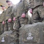 Группа молодых людей в камуфляжной форме стоит в помещении рядом с армейскими вещмешками, на сумках видна надпись «АРМИЯ РОССИИ».