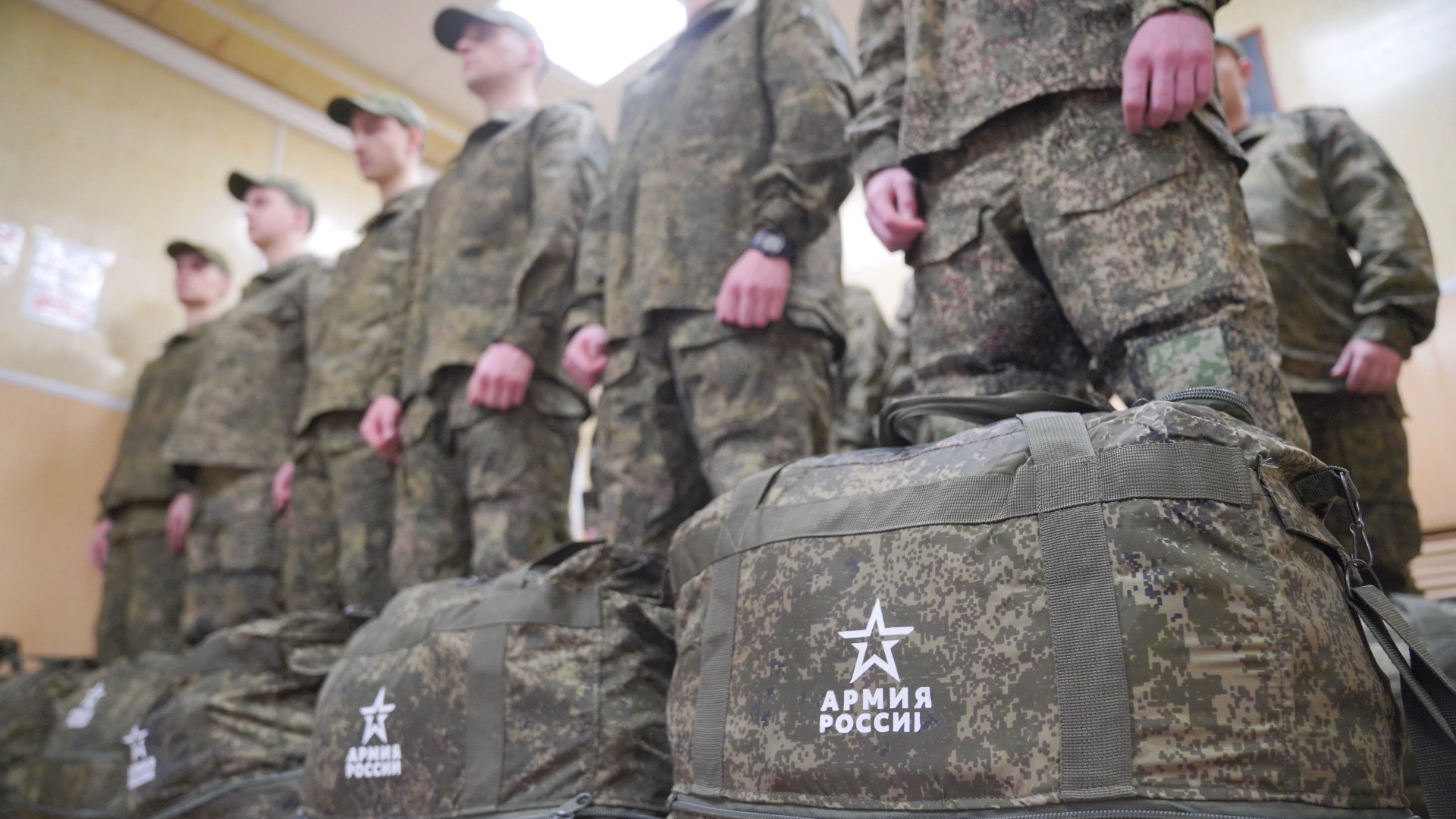 Группа молодых людей в камуфляжной форме стоит в помещении рядом с армейскими вещмешками, на сумках видна надпись «АРМИЯ РОССИИ».