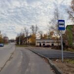 Уличный кадр: дорожное полотно, знак автобусной остановки и недавно обустроенная посадочная площадка у жилой застройки; автомобили и осенние деревья.