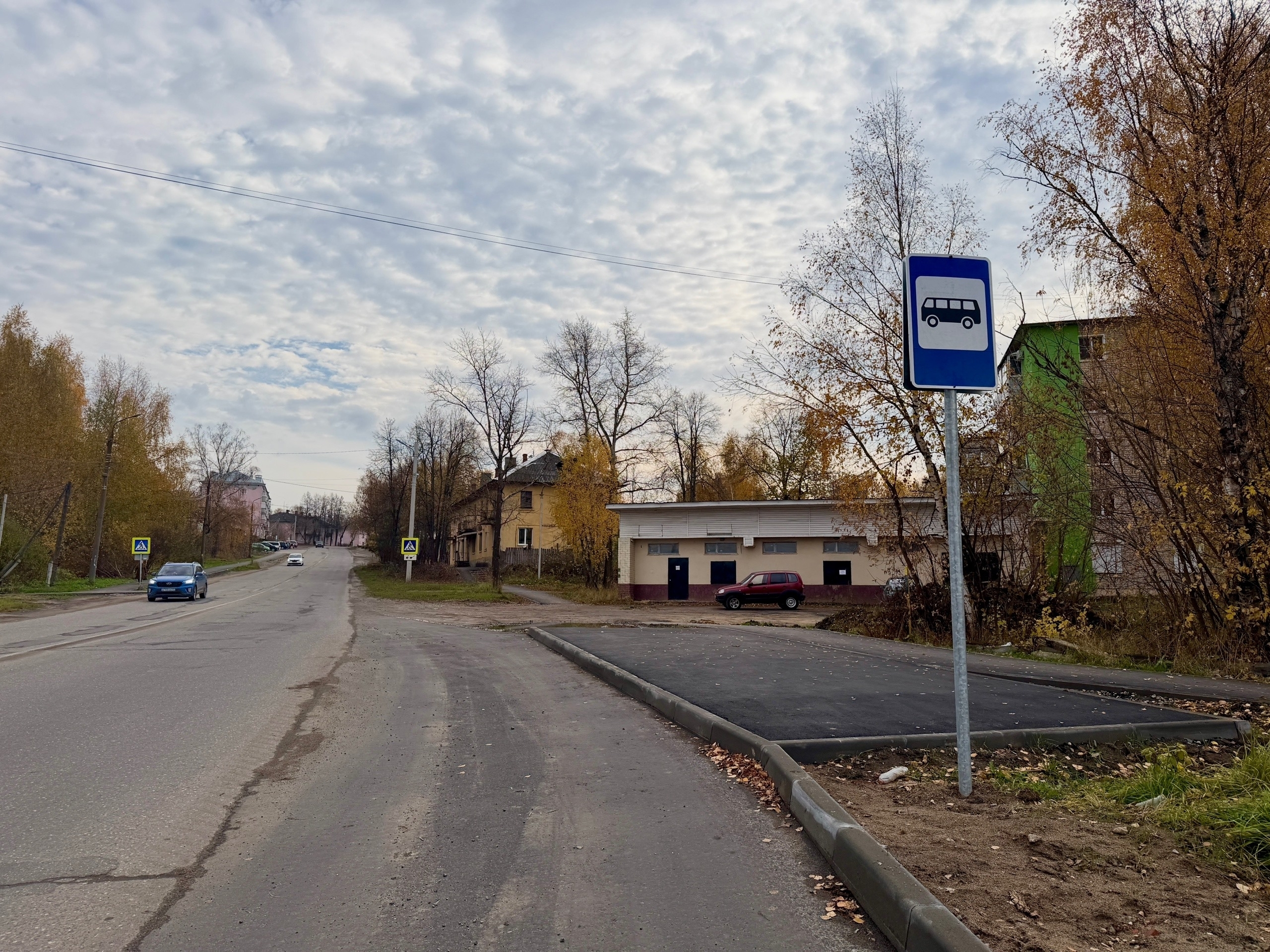 Уличный кадр: дорожное полотно, знак автобусной остановки и недавно обустроенная посадочная площадка у жилой застройки; автомобили и осенние деревья.
