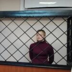 Женщина в тёмно-бордовом свитере стоит за металлической решёткой (клеткой для подсудимых) в зале суда