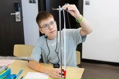 школьник за партой выполняет физический физический эксперимент на измерительной стойке с подвешенным грузом
