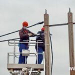 Два электромонтажника в касках и спецодежде на подъёмнике обслуживают провода линии электропередачи.