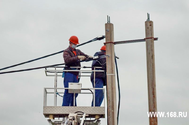 Два электромонтажника в касках и спецодежде на подъёмнике обслуживают провода линии электропередачи.