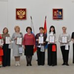 Группа женщин в белом зале держит рамки с грамотами и подарочные пакеты, на стене — гербы и флаги; сценa награждения.