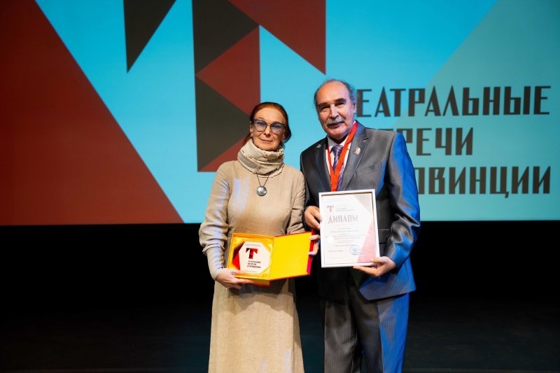 Двое человека на сцене держат открытую коробку с наградой и бумажный диплом; на заднем плане экран с надписью «Театральные встречи в провинции».