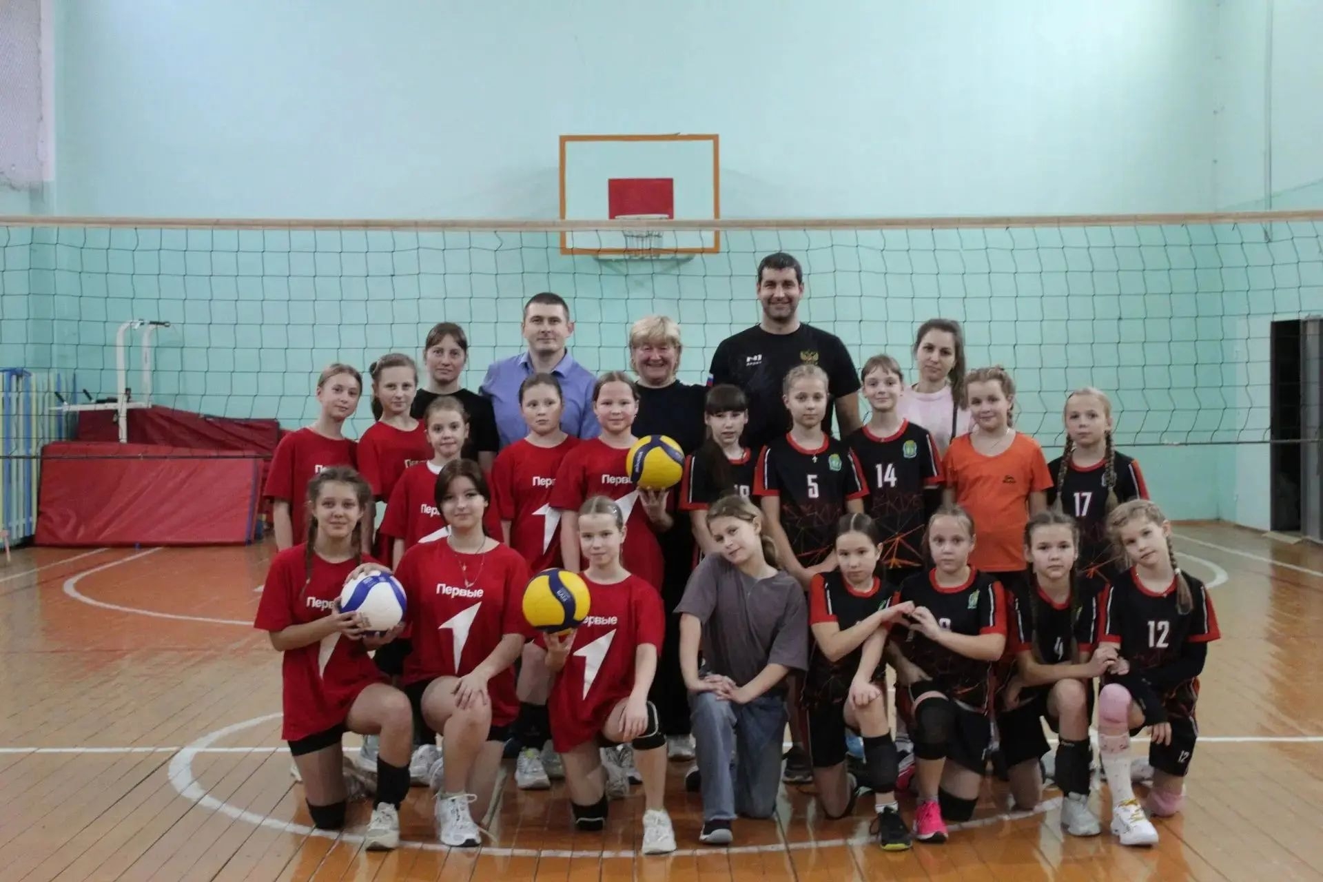 Групповое фото юношеских волейбольных команд и тренеров в спортивном зале у сетки; девушки в красной и чёрной форме держат мячи