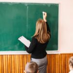 Учительница стоит спиной и пишет мелом на зелёной школьной доске; на переднем плане видны головы учеников.
