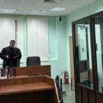 зал судебного заседания: судья у скамьи, женщина у стола, стеклянная клетка для подсудимых и присутствующий мужчина