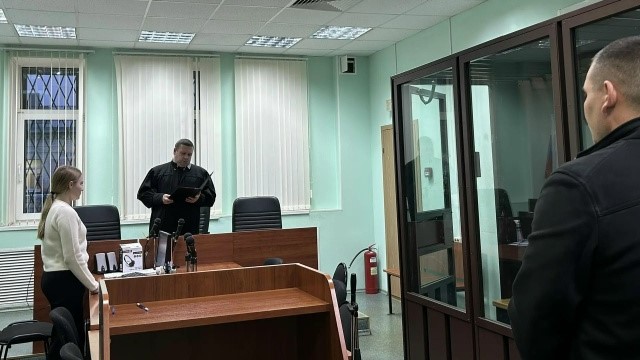 зал судебного заседания: судья у скамьи, женщина у стола, стеклянная клетка для подсудимых и присутствующий мужчина