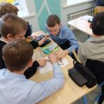 Группа школьников за парой в классе, мальчики рассматривают и обсуждают распечатку с силуэтами животных; на столах пеналы, линейка и другие школьные принадлежности.