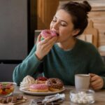 женщина за кухонным столом ест пончик; на столе тарелка с пончиками, зефир, кусочки шоколада и миска с мармеладом, чашка напитка