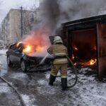 пожарный в спецформе поливает из рукава горящий легковой автомобиль рядом с горящим гаражом на заснечной улице; пожарная машина на заднем плане
