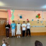 Группа школьников на фоне украшенной стены с воздушными шарами, бумажными вырезками персонажей и большими цифрами; сцена в школьном коридоре/классе.