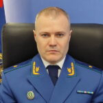 Портрет мужчины в синей прокурорской форме за креслом на фоне официального флага и эмблемы