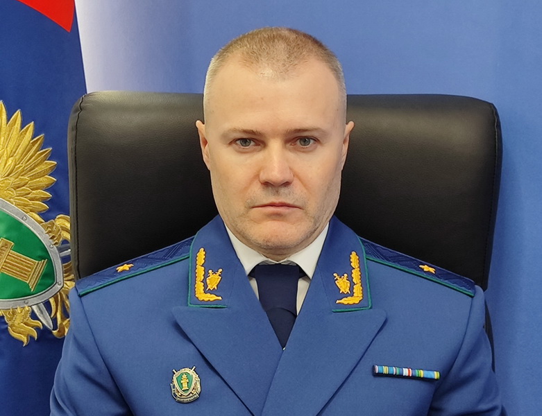 Портрет мужчины в синей прокурорской форме за креслом на фоне официального флага и эмблемы