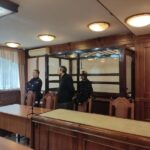Судебный зал с металлической кабиной для подсудимых; мужчина в гражданской одежде и сотрудник полиции у кабины; столы и кресла в зале.