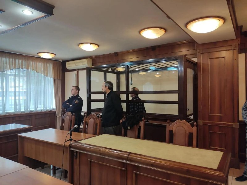 Судебный зал с металлической кабиной для подсудимых; мужчина в гражданской одежде и сотрудник полиции у кабины; столы и кресла в зале.