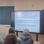 Студенты в аудитории (спинами) смотрят на большой экран с презентацией в классе.