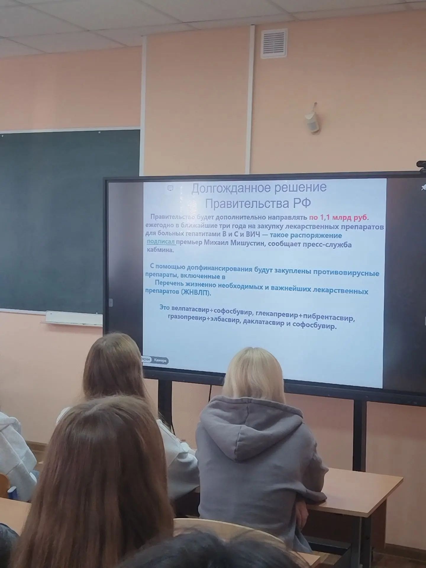 Студенты в аудитории (спинами) смотрят на большой экран с презентацией в классе.