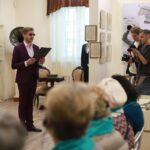 мужчина в костюме читает с папкой в экспозиционном зале музея; перед ним сидящая аудитория, справа — фотографы и музейная карта на стене