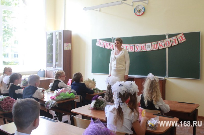 Учительница у школьной доски, первоклассники/ученики начальных классов за партами с букетами