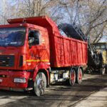 Фрезеровочная машина снимает верхний слой дорожного покрытия; рядом грузовой самосвал красного цвета и рабочие на проезжей части.