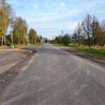 Сельская асфальтированная улица с новыми слоями покрытия, гравийными обочинами, деревьями и домами по сторонам