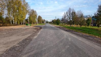 Сельская асфальтированная улица с новыми слоями покрытия, гравийными обочинами, деревьями и домами по сторонам