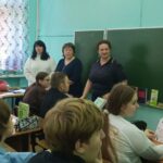 Ученики за партами в классе; у доски стоят три взрослые (одна в форме), школьная доска, учебники на столах.