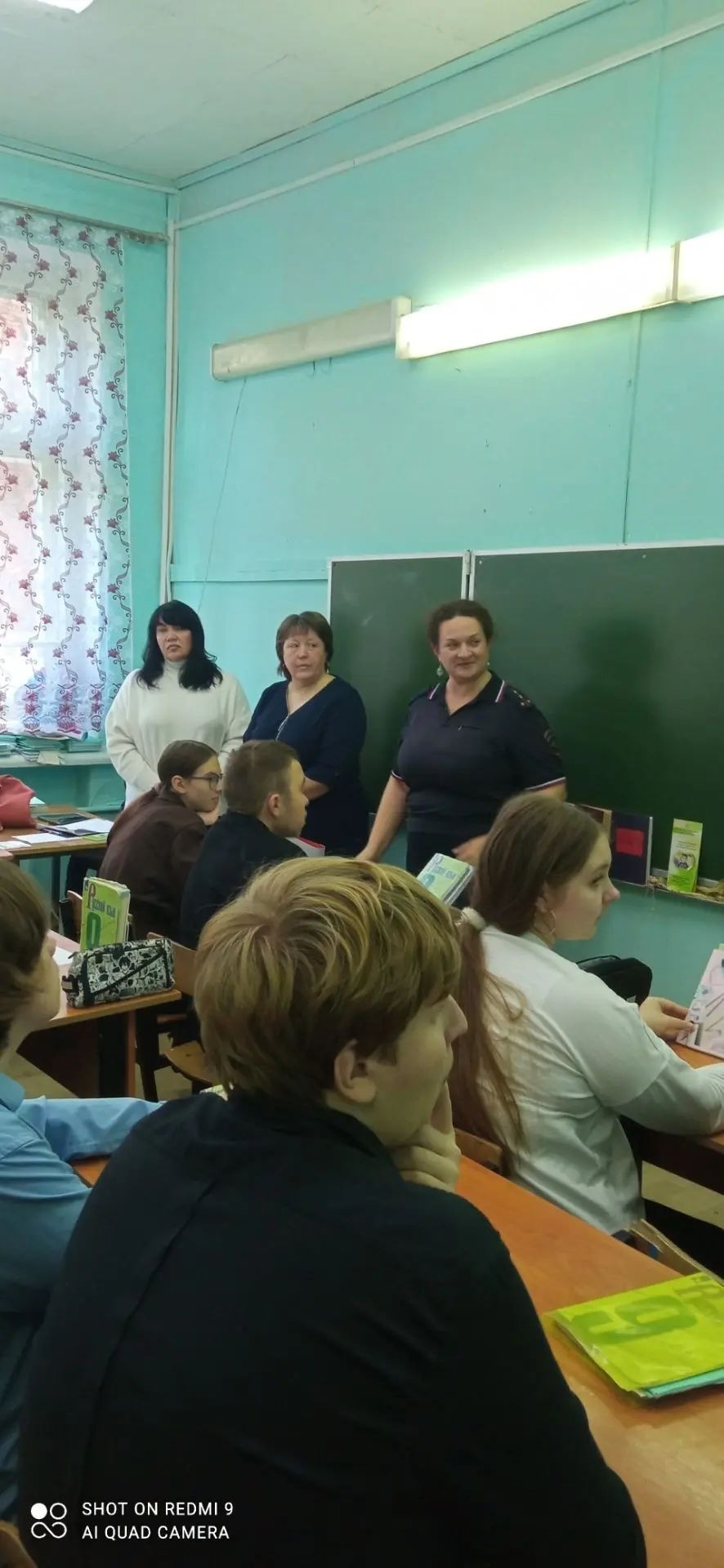 Ученики за партами в классе; у доски стоят три взрослые (одна в форме), школьная доска, учебники на столах.