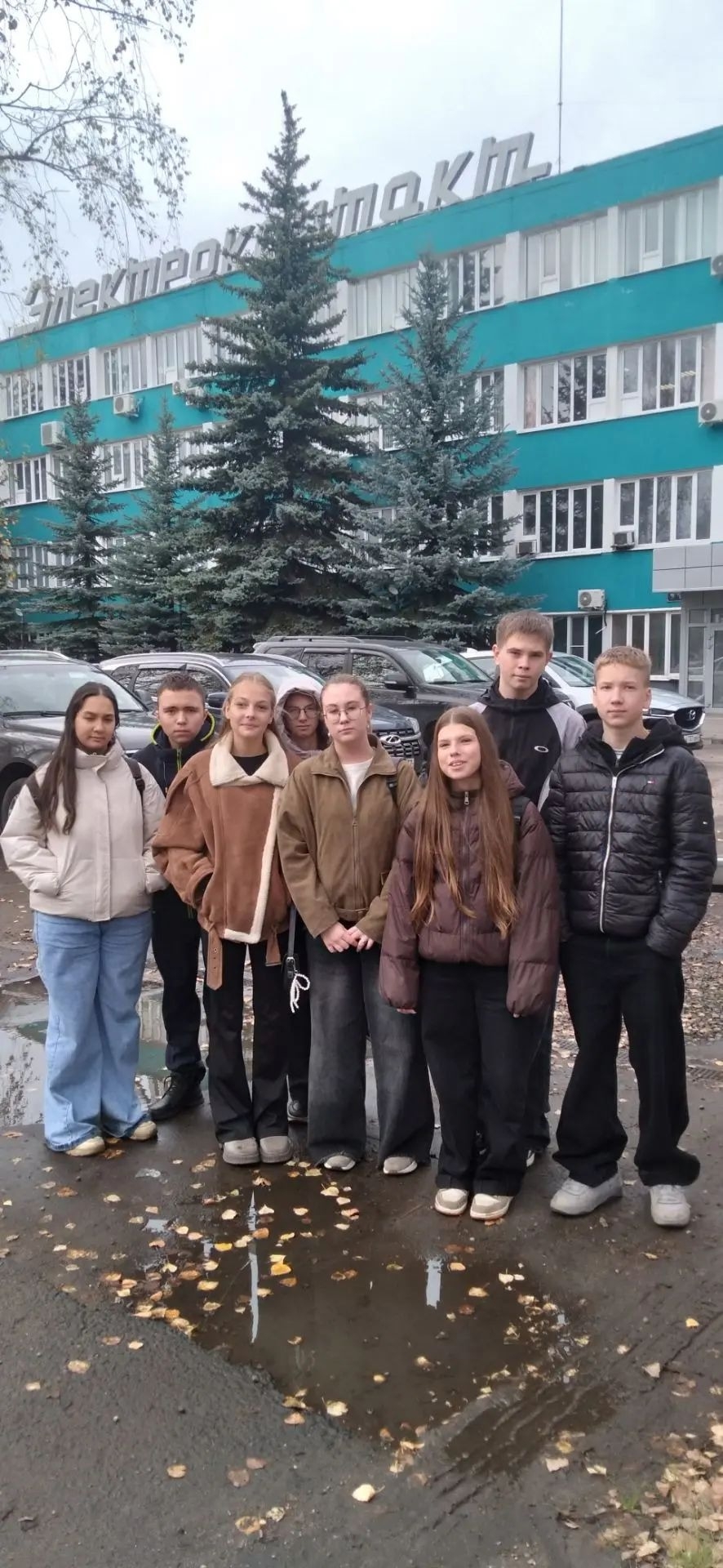 Группа школьников стоит на улице перед зелёным заводским зданием с крупной вывеской «Электроконтакт».
